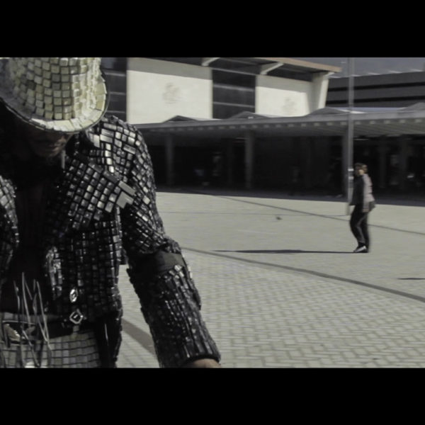Video still in front of Cape Town Train station, with African contemporary artist Maurice Mbikayi dressed in his keyboard clothing